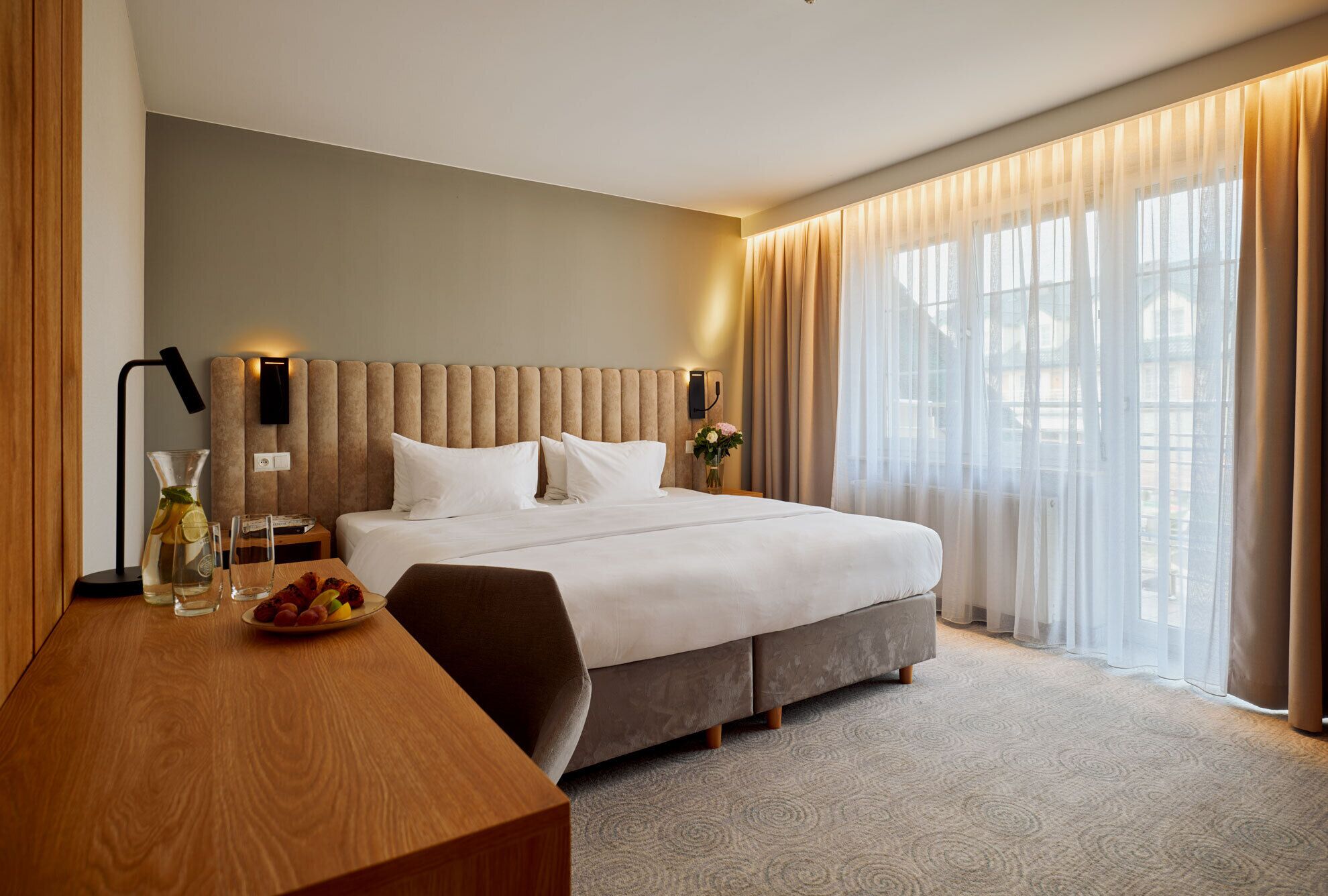 A modern hotel room with a large bed, a wooden desk, and a large window with sheer curtains.