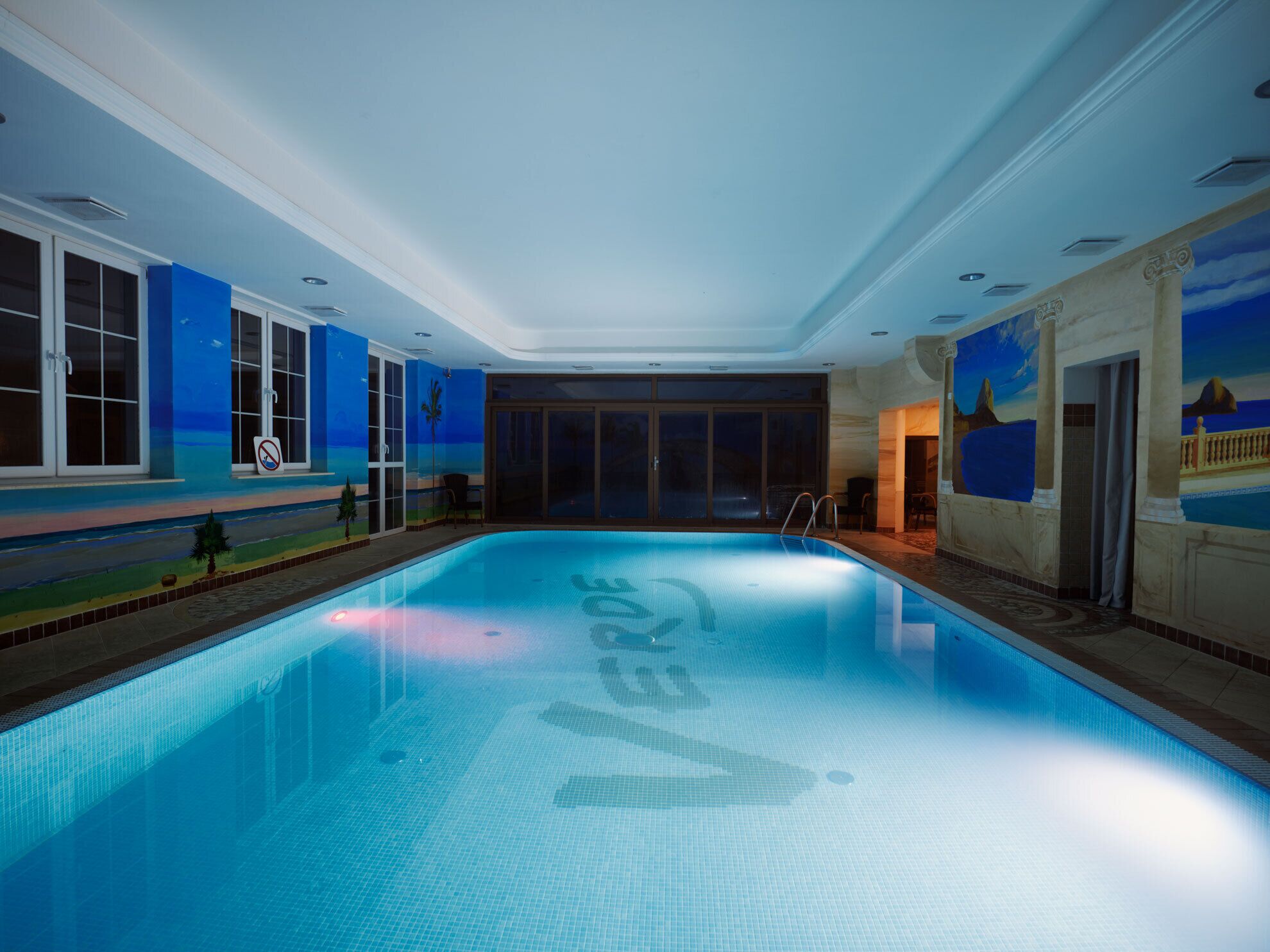 An indoor swimming pool with clear blue water, surrounded by walls with a mural of a beach scene.