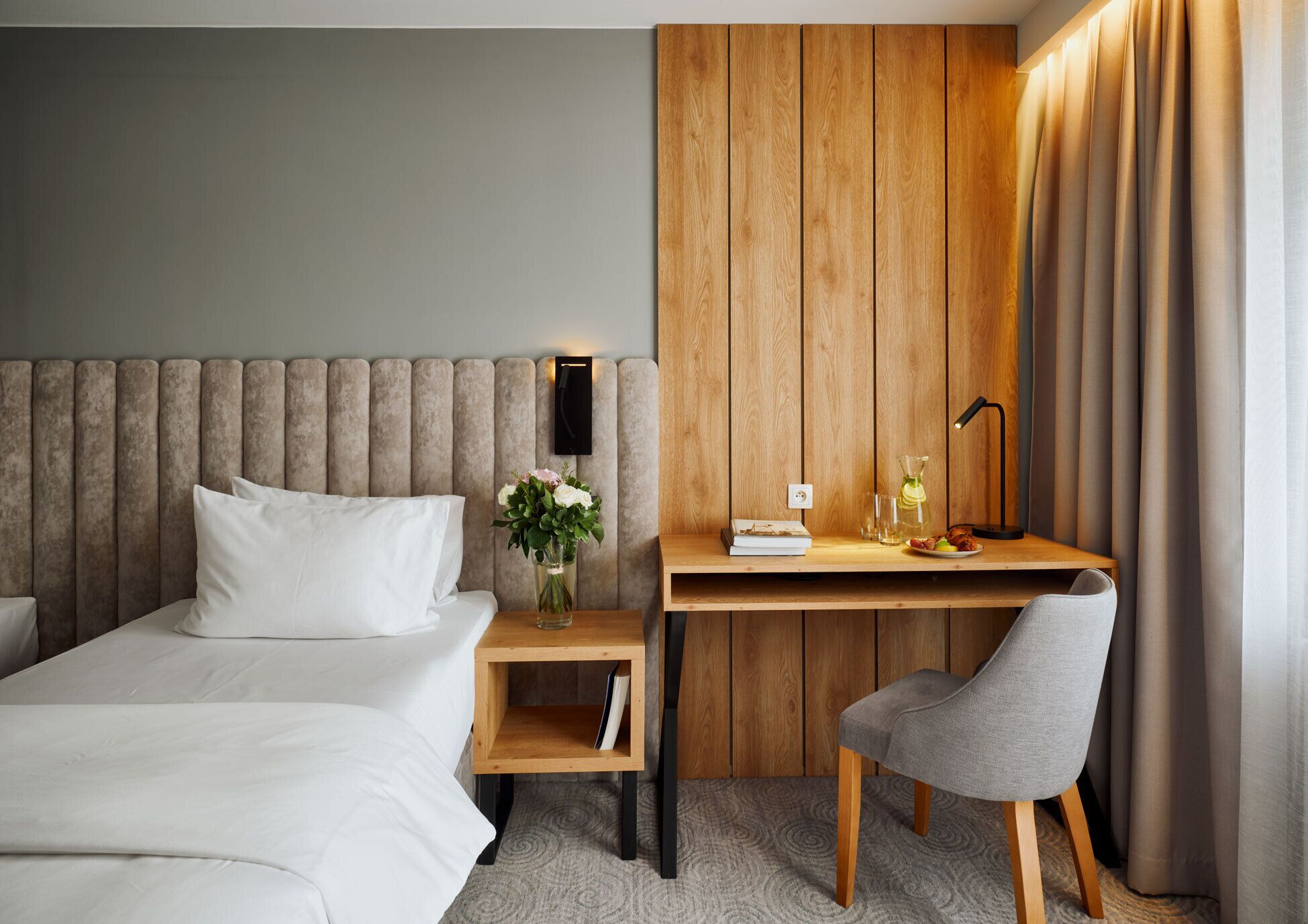 A hotel room with a bed, a desk with a chair, a vase with flowers, and a lamp.