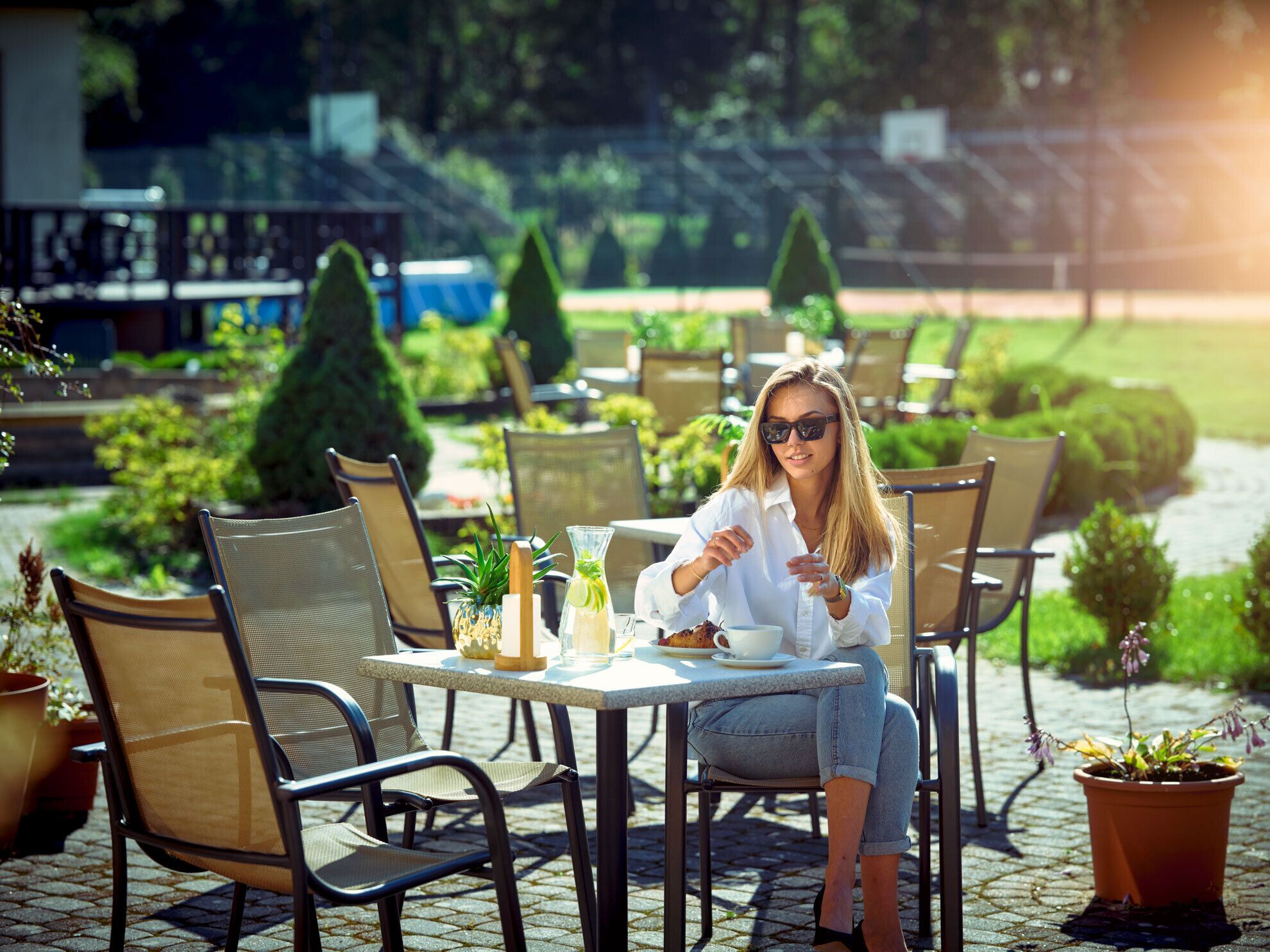 A woman seated at an outdoor cafe with a table, chairs, and potted plants.