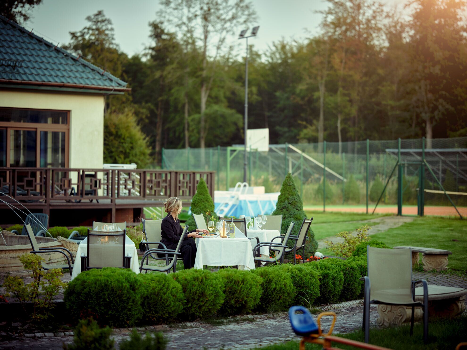 Un espace de restauration en plein air, avec des tables et des chaises, entouré de haies bien entretenues et agrémenté d’un petit élément décoratif aquatique.
