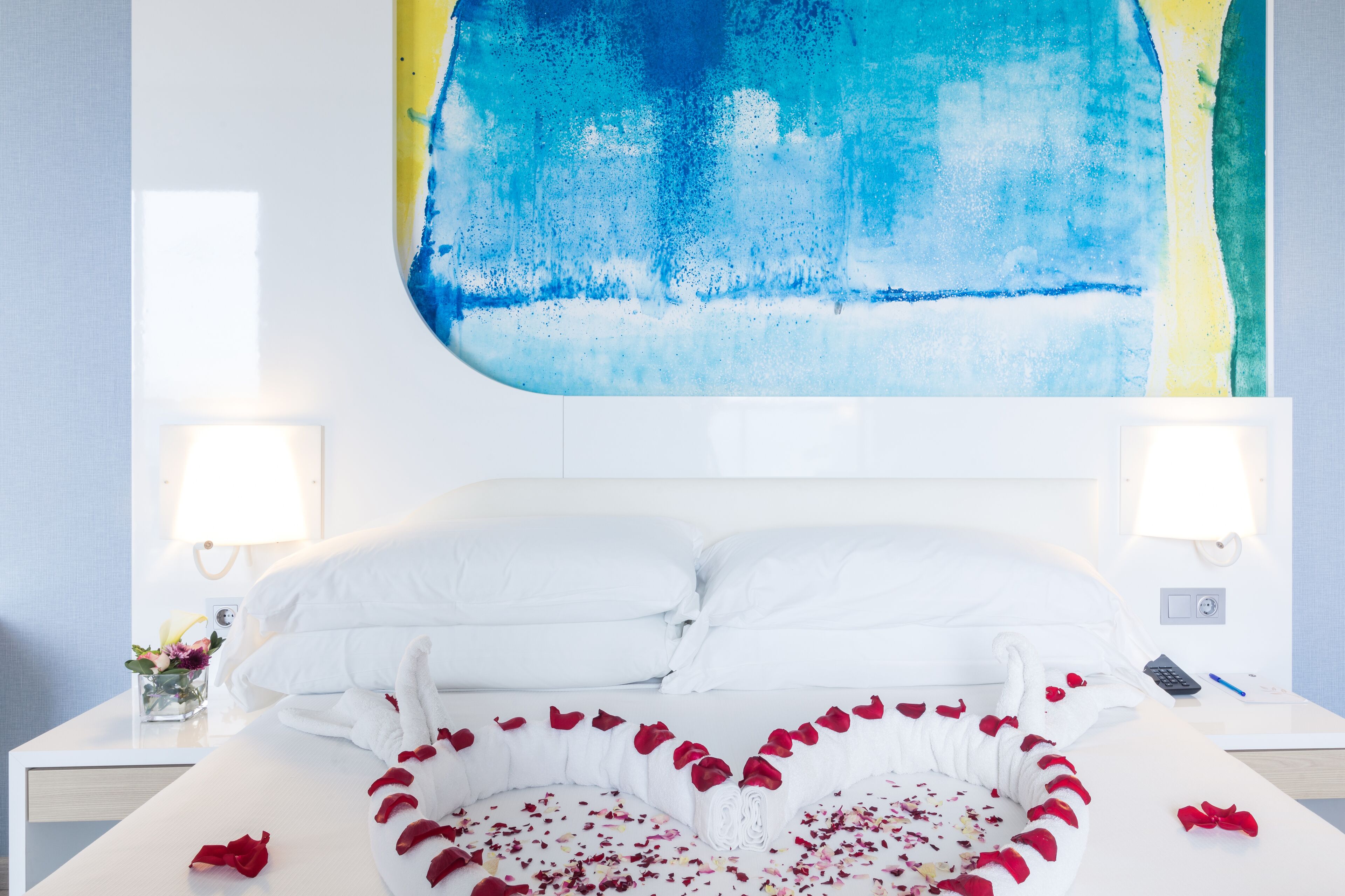 Habitación de hotel con cama, ropa de cama blanca, un arreglo de pétalos de rosa en forma de corazón y un cuadro abstracto grande en la pared.