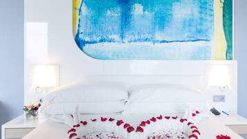 Habitación de hotel con cama, ropa de cama blanca, un arreglo de pétalos de rosa en forma de corazón y un cuadro abstracto grande en la pared.