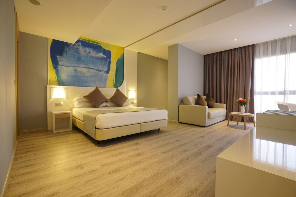 Ein modernes Hotelzimmer mit einem großen Bett, einer Couch, einem kleinen Tisch mit einer Vase und einem großen abstrakten Gemälde an der Wand.