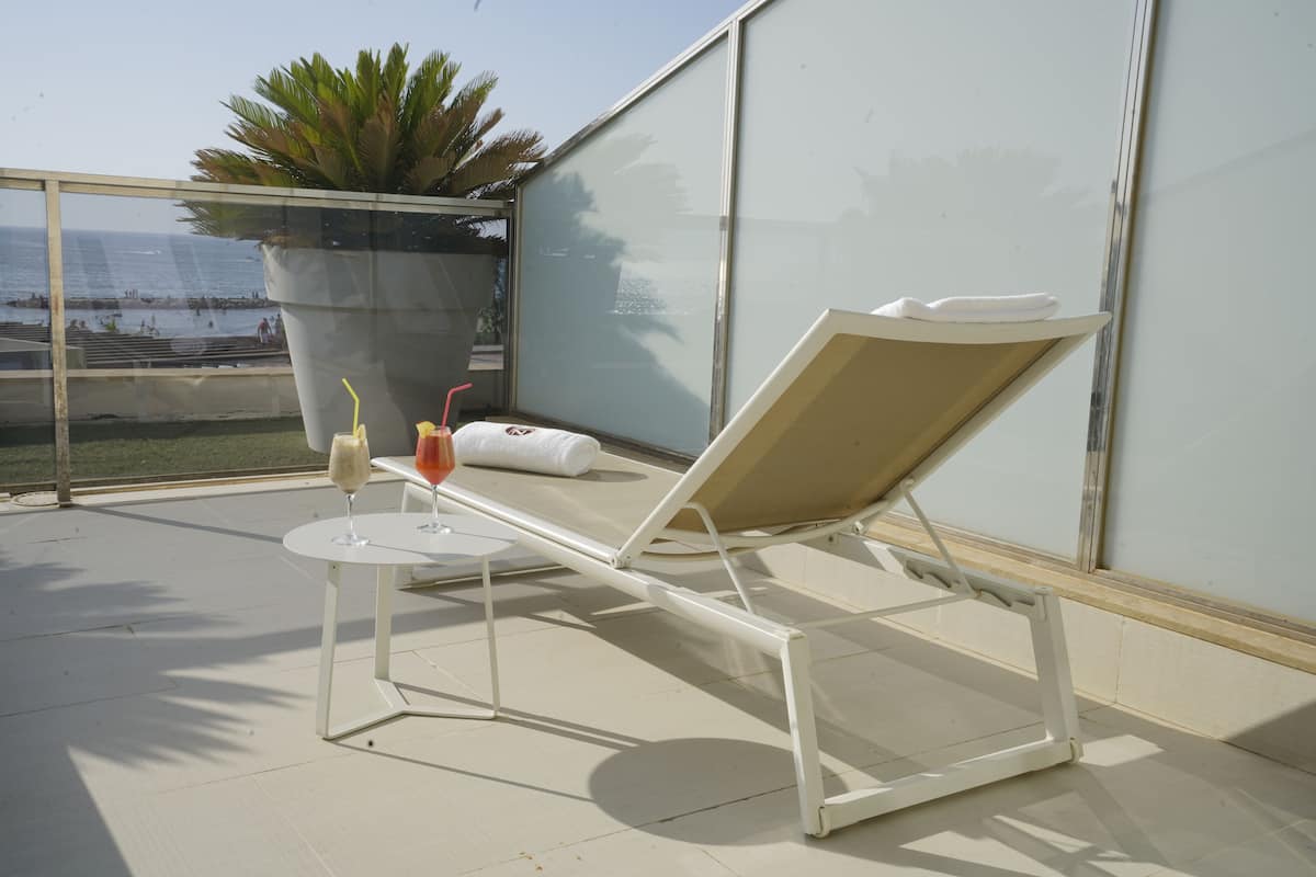 Ein sonniger Balkon mit Liegestuhl, einem kleinen Tisch mit Getränken und einem großen Topfpflanzen.