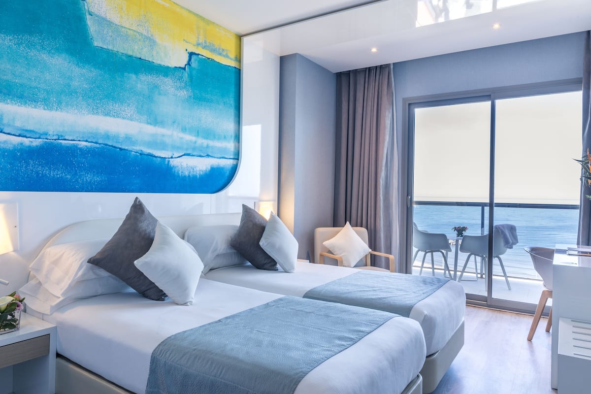 Ein modernes Hotelzimmer mit zwei Betten, einem großen Fenster mit Meerblick, einem Schreibtisch mit Stuhl und einem Gemälde an der Wand.