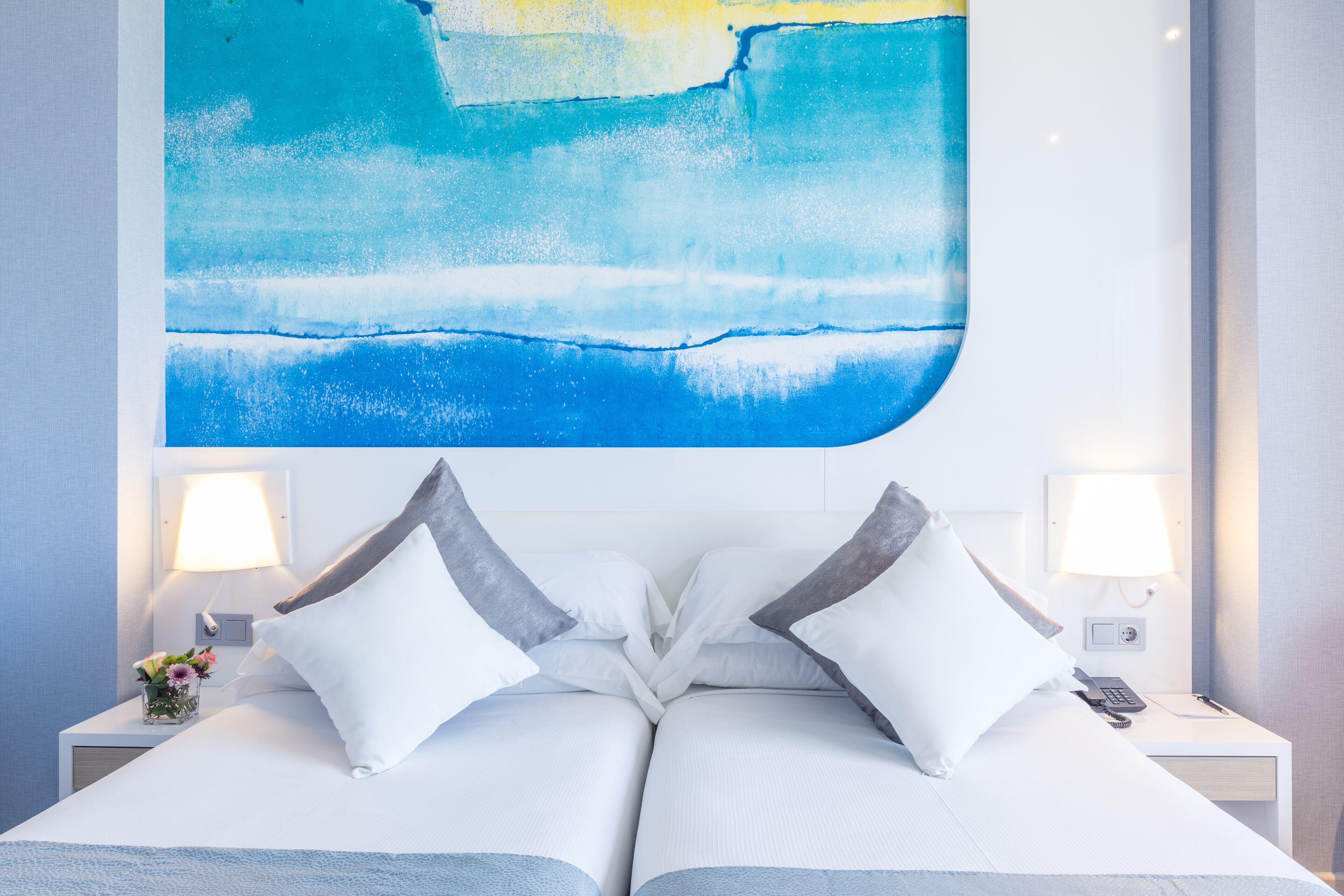 A hotel room with a large abstract painting of blue and yellow hues on the wall, a neatly made bed with white and gray pillows, two bedside tables with lamps, and a small vase with flowers.