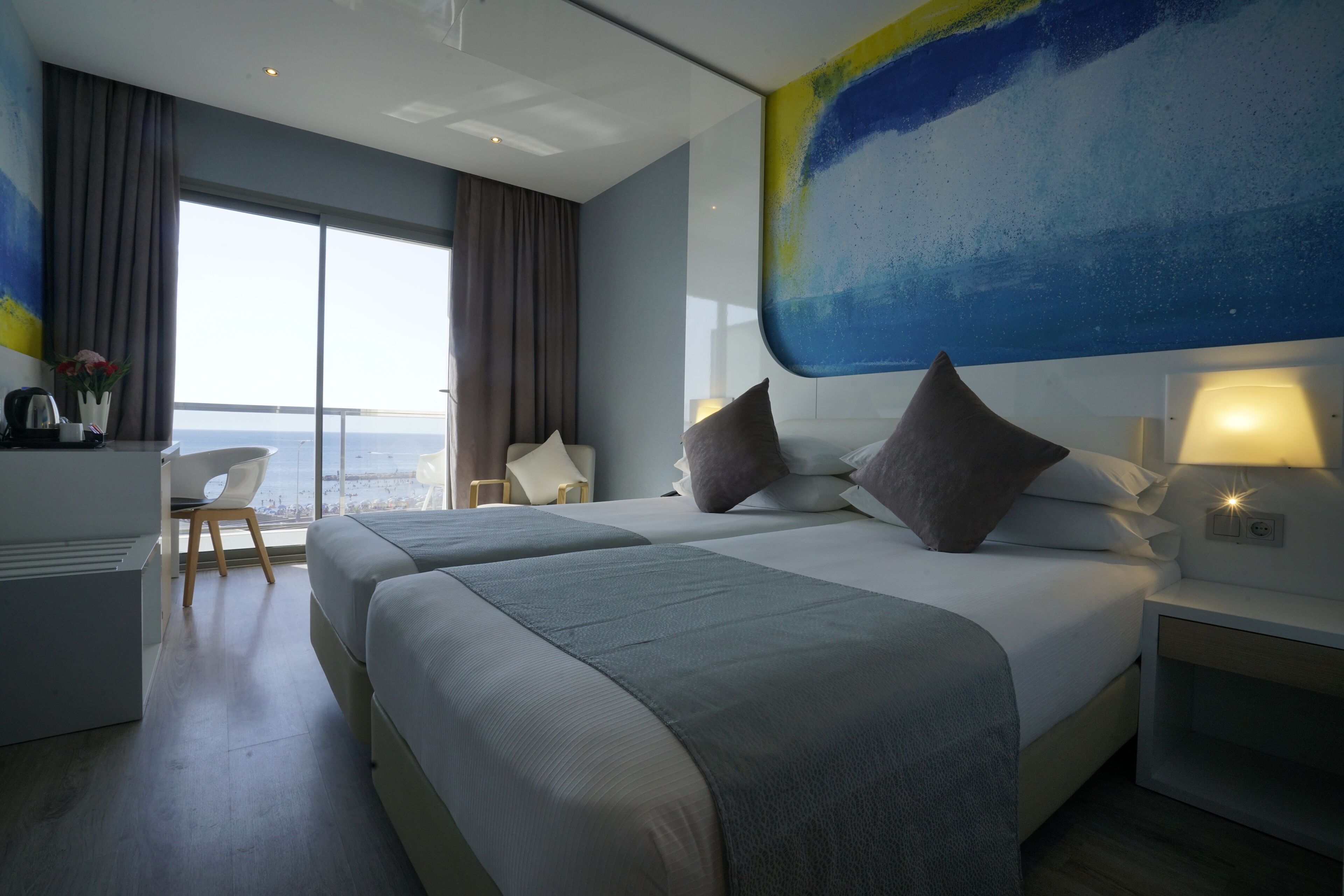 Habitación de hotel moderna con dos camas, un escritorio y un amplio ventanal con vista al mar.