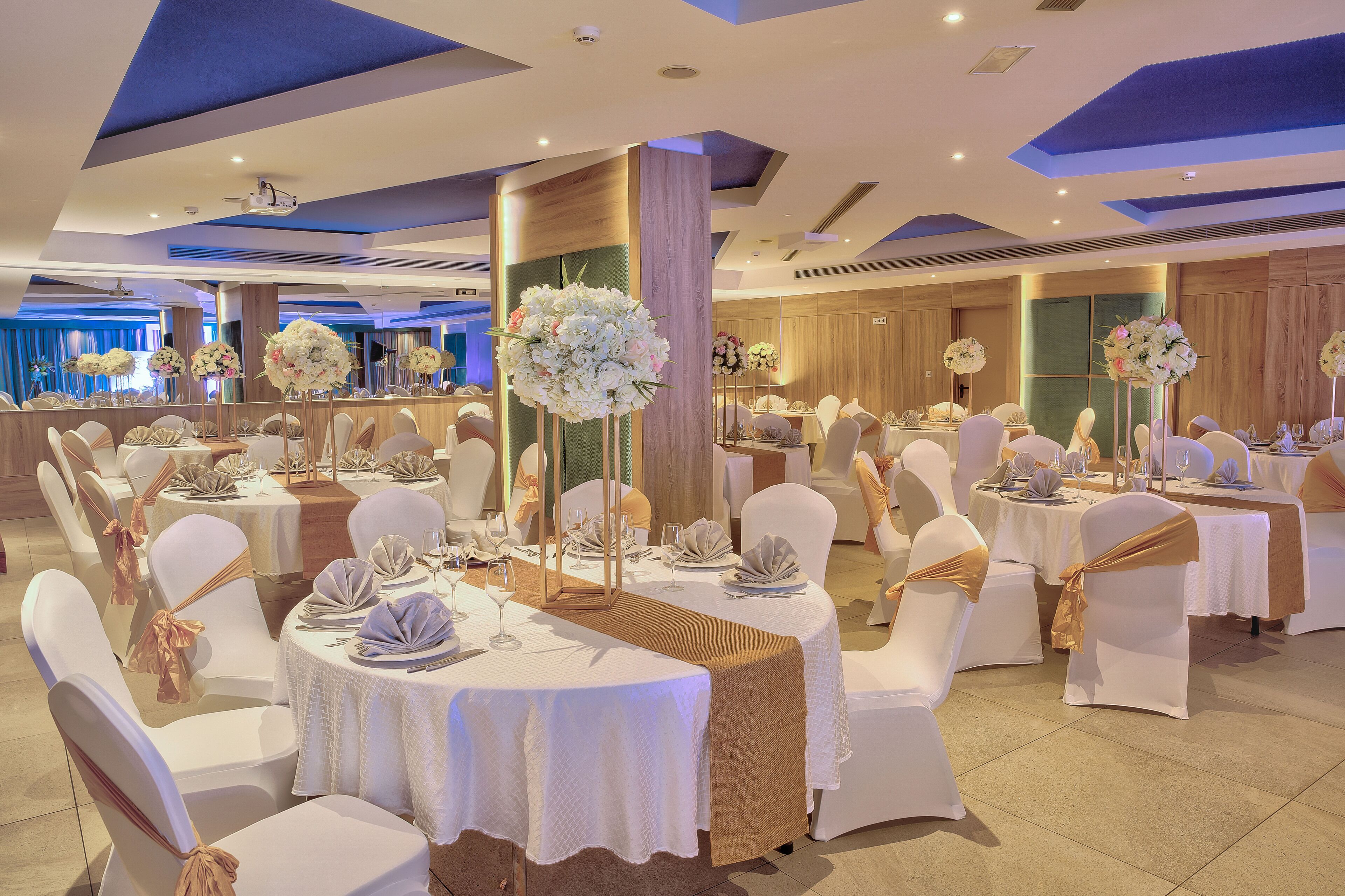 Une salle de banquet avec des tables rondes, des chaises blanches et des centres de table ornés de compositions florales.