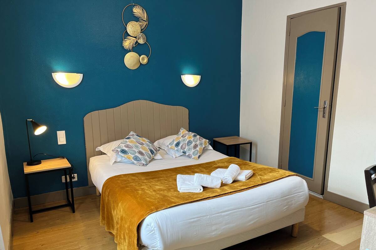 Una cama bien hecha con una manta marrón, dos mesitas de noche con lámparas, almohadas estampadas y un adorno en la pared.