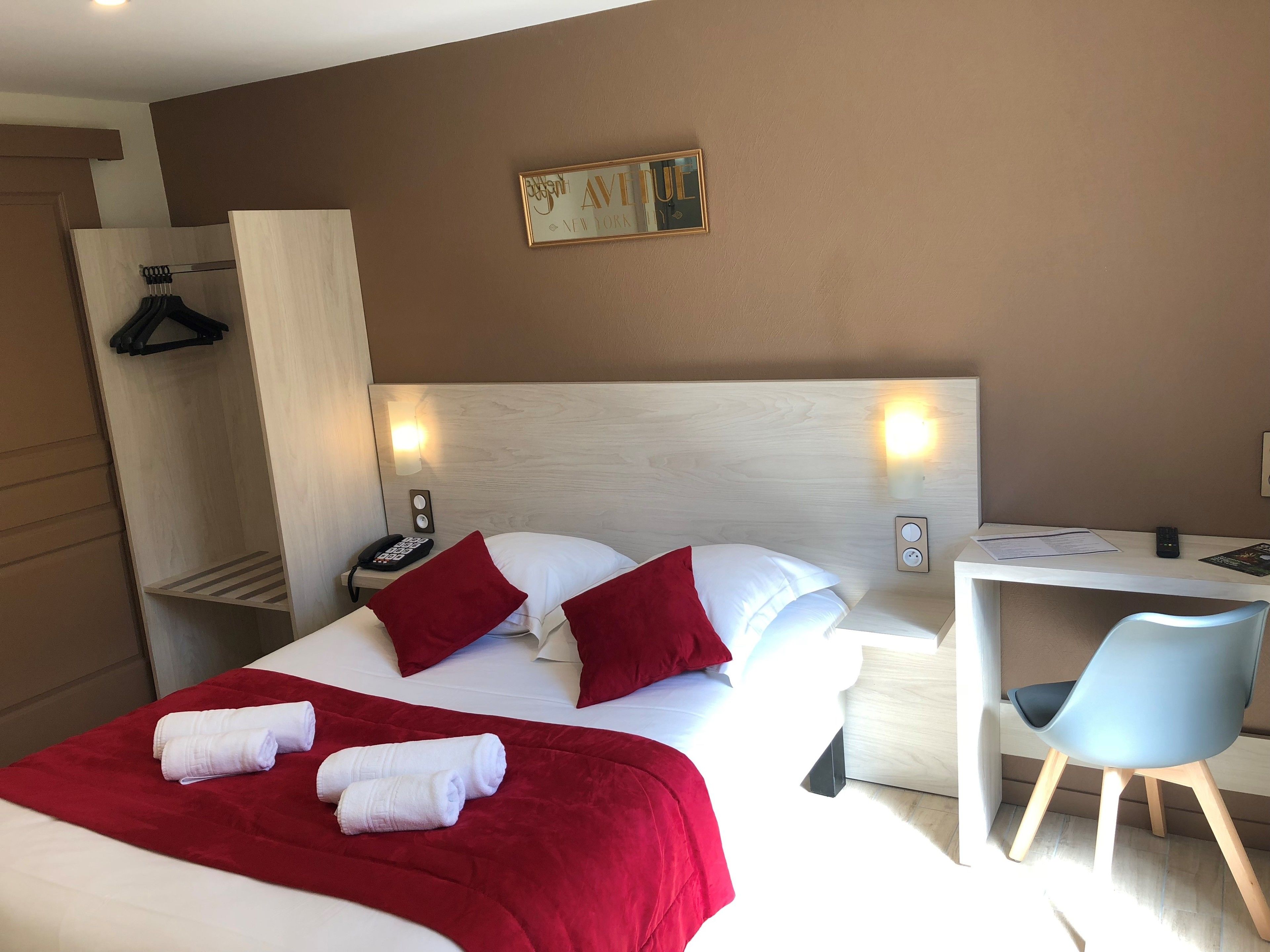 A hotel room with a bed, two bedside tables, a desk, a chair, a hanging coat rack, and a wall-mounted artwork.