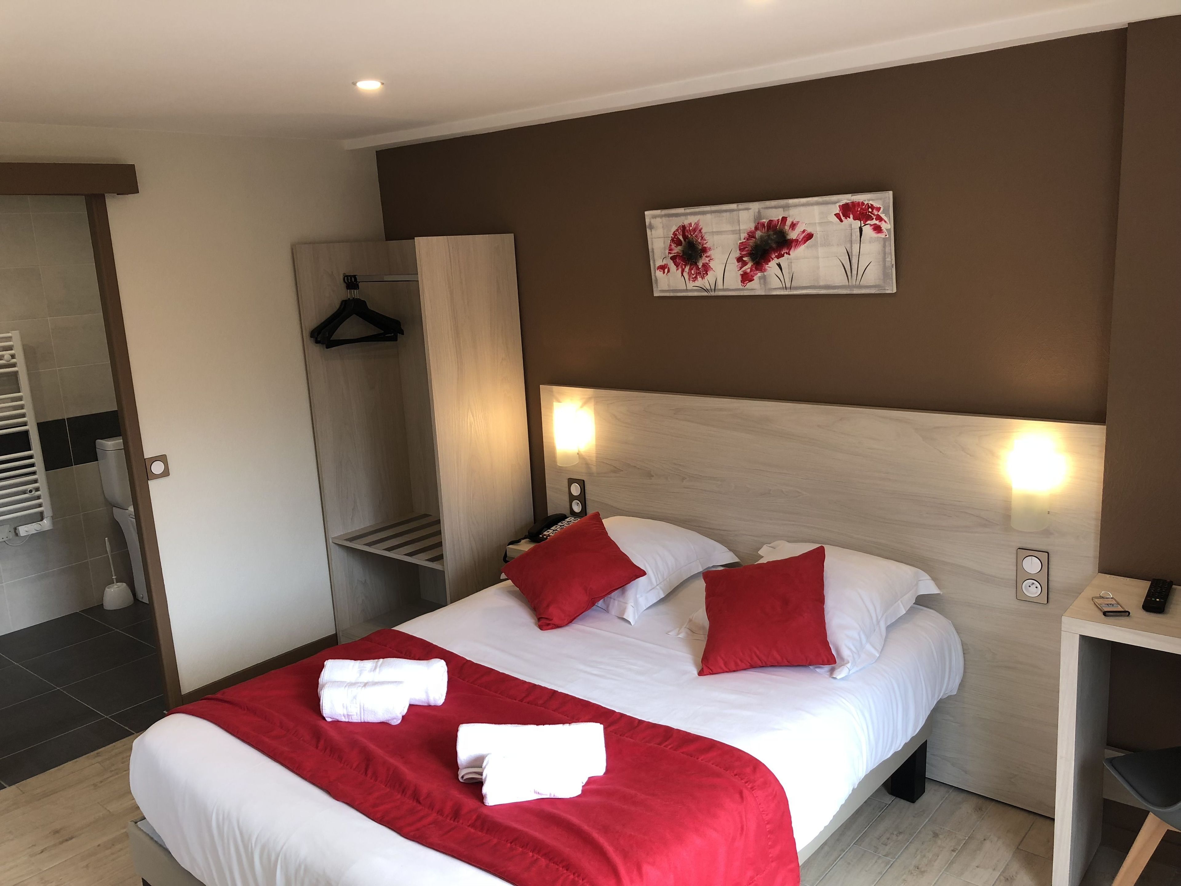 Habitació d'hotel amb llit, roba de llit vermella i blanca, un capçal de llit de fusta, un quadre de flors, una taula de nit amb un telèfon i una làmpada, i un penjador de tovalloles.