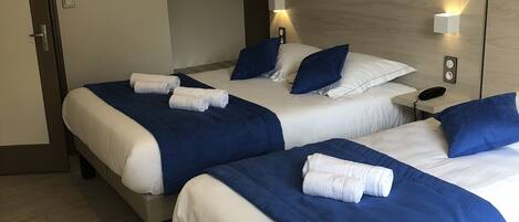 Habitación de hotel con dos camas, cada una con ropa de cama azul y blanca, un cuadro de veleros en la pared y una mesita de noche con un control remoto.