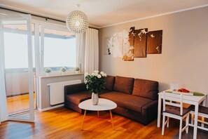 Un salon moderne avec un canapé marron, une table basse blanche et une table à manger blanche avec des chaises. Une grande fenêtre avec des rideaux, un radiateur et un pot de fleurs sont présents sur le rebord de la fenêtre.