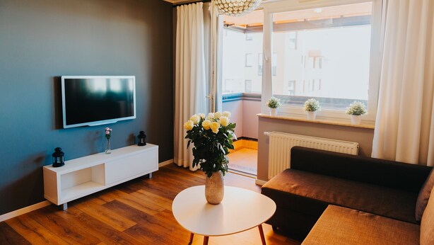 A modern living room with a flat-screen TV, a white console table, a round white table, a brown sofa, and a large window with curtains. - Elite Apartments Seaview Horyzont (Gdansk)