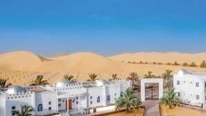 Un resort en el desierto con edificios blancos, palmeras y dunas de arena al fondo.