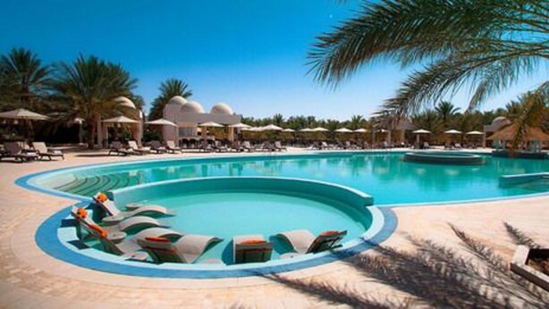 Una gran piscina al aire libre con sillones reclinables y sombrillas, rodeada de palmeras y zonas de sombra.