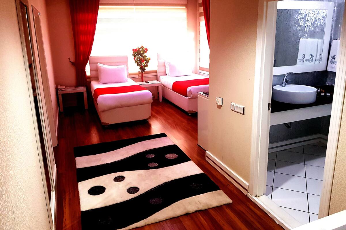A hotel room with a bed, a nightstand, a desk, a bathroom with a sink, and a window with curtains.