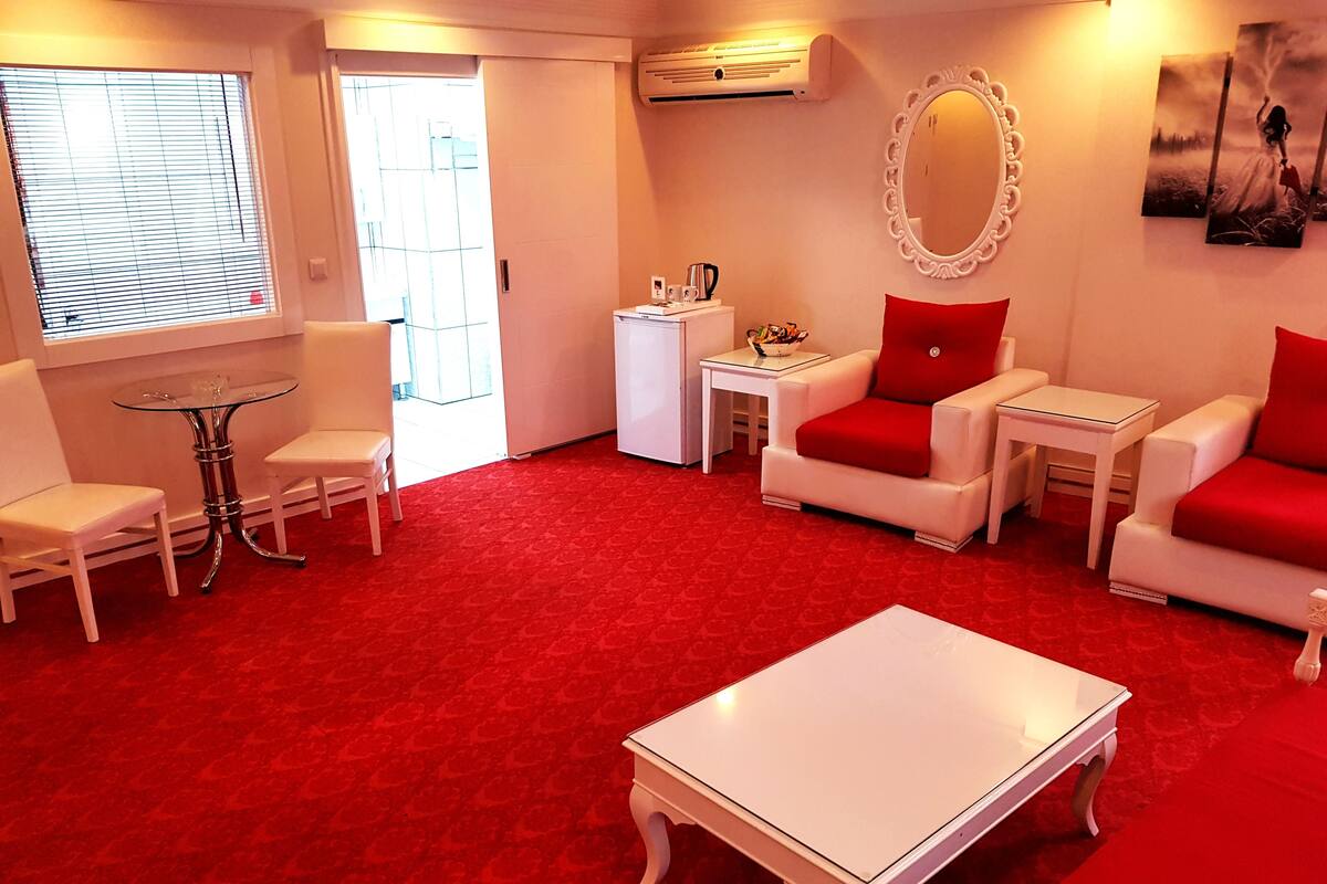 A living room with a red carpet, white furniture, a glass coffee table, a white sofa with red cushions, a white armchair, a small white side table, and a white cabinet.