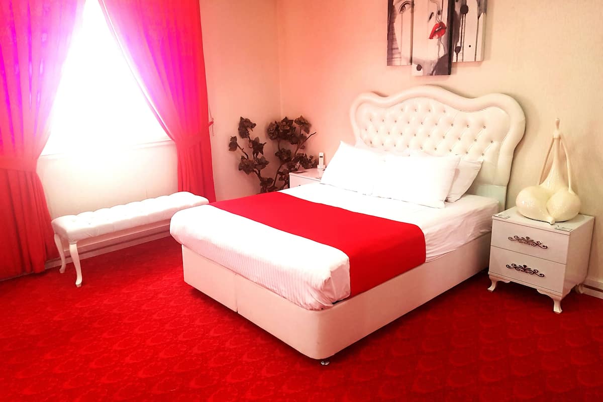 A bedroom with a white bed, red accents, a bench, a nightstand, and a wall art piece.