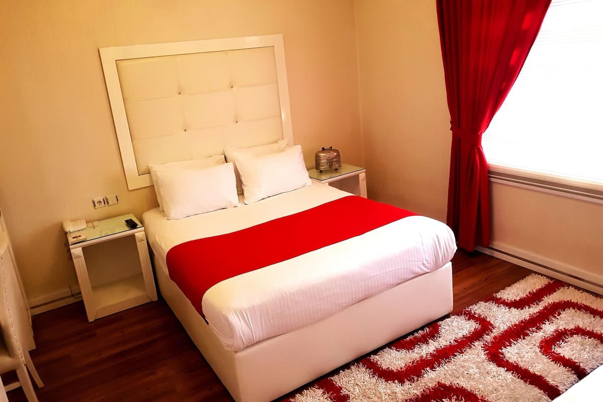 A neatly made bed with a red and white bedspread, a white headboard, and a bedside table with a lamp and a small item on it.