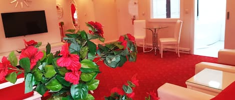 Una sala de estar con alfombra roja, muebles blancos, un televisor de pantalla plana y una planta artificial grande.