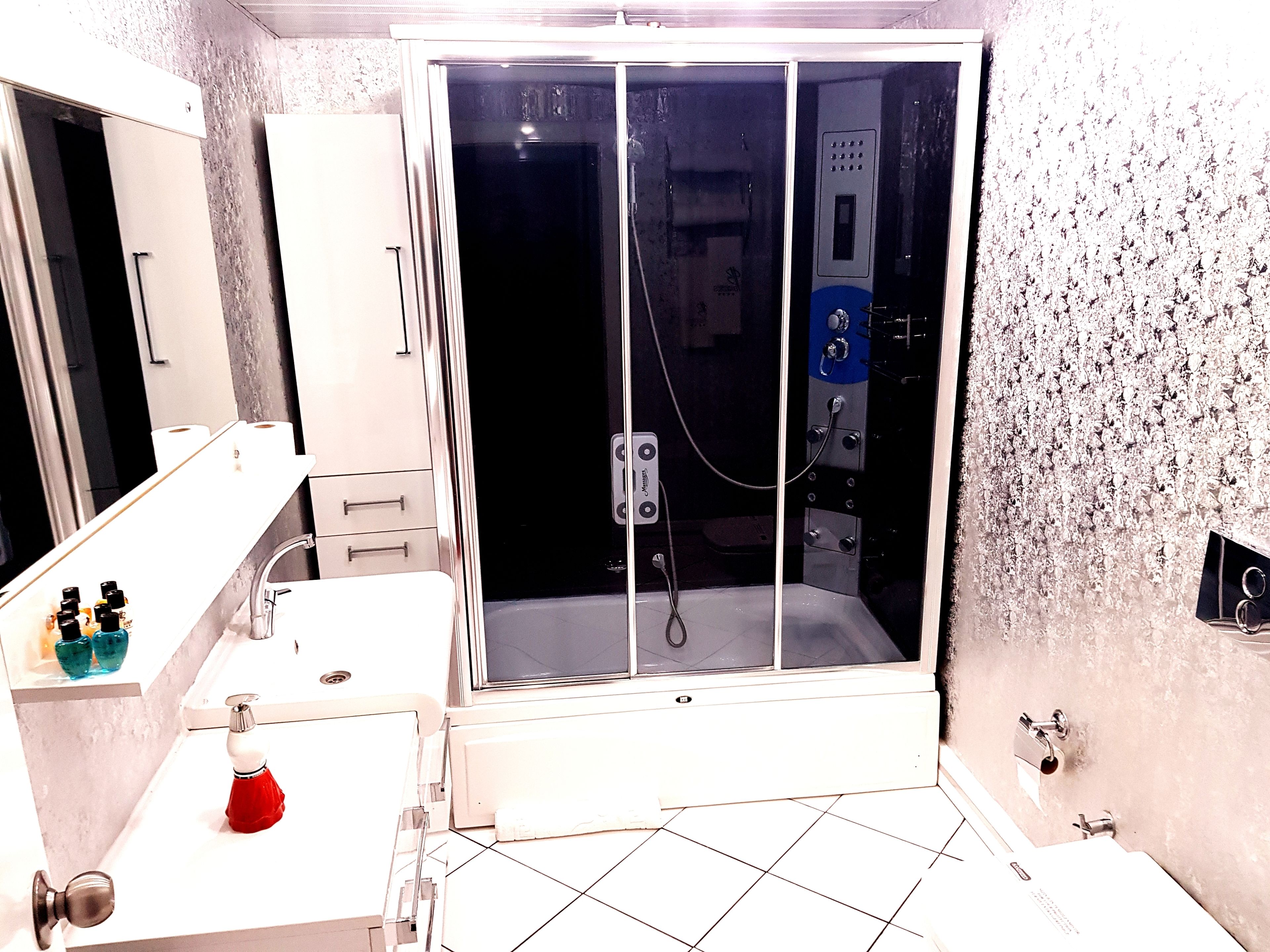 A bathroom with a glass-enclosed shower, a white sink with a red soap dispenser, and a mirror above the sink.
