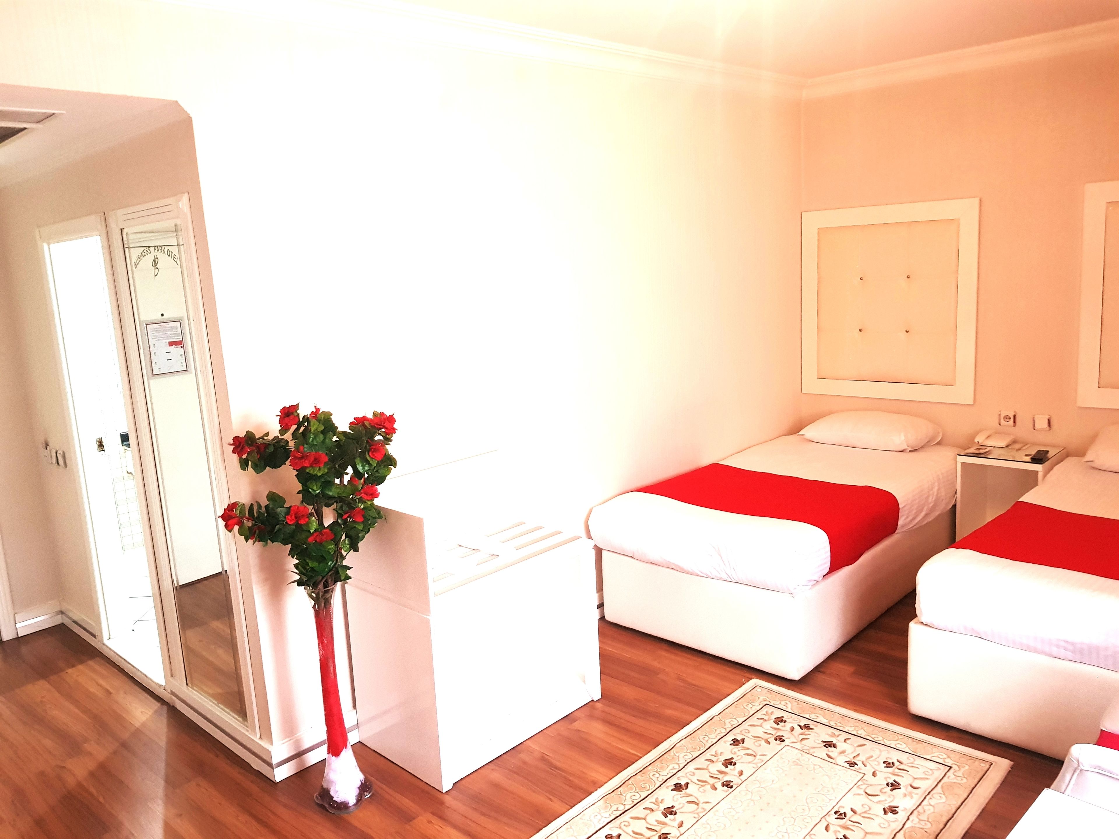 A hotel room with two beds, a small desk, a vase with red flowers, and a wooden floor.