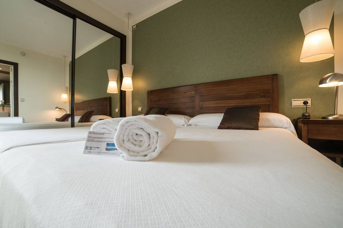 Een keurig opgemaakt bed met een opgerolde witte handdoek en een label, een houten hoofdbord en twee bedlampjes.