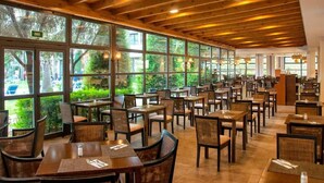 Een ruime, overdekte eethoek met houten balken in het plafond, grote ramen met uitzicht op groen, en meerdere tafels en stoelen klaar voor een maaltijd.