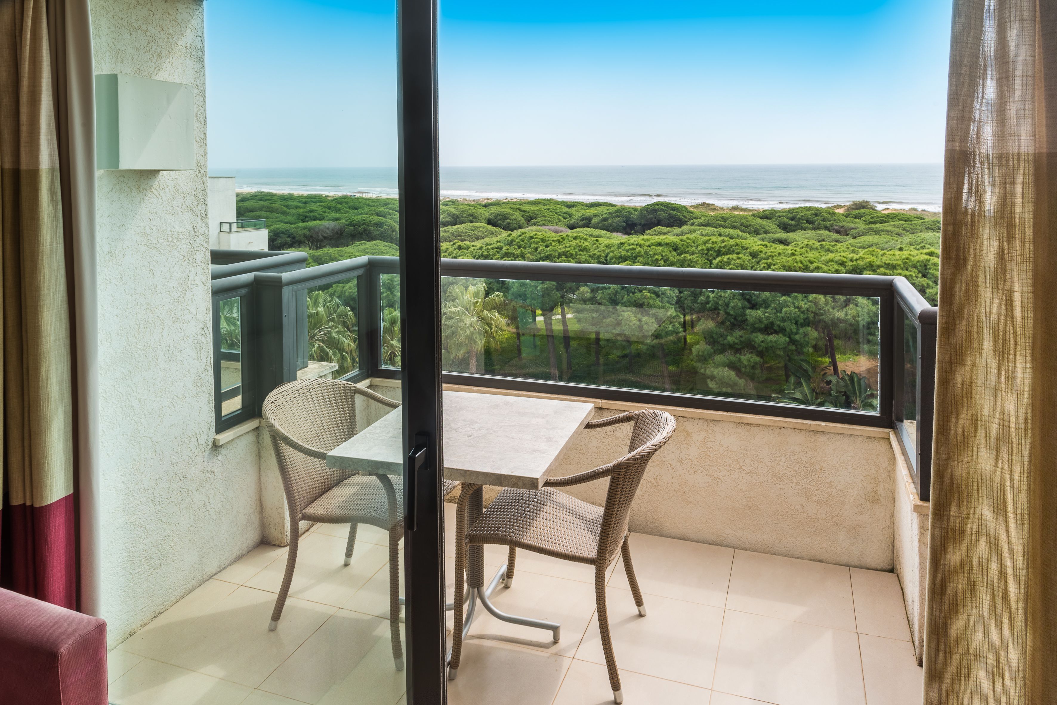 Un balcón con mesa y sillas, con vista a un paisaje costero con vegetación densa y el océano.
