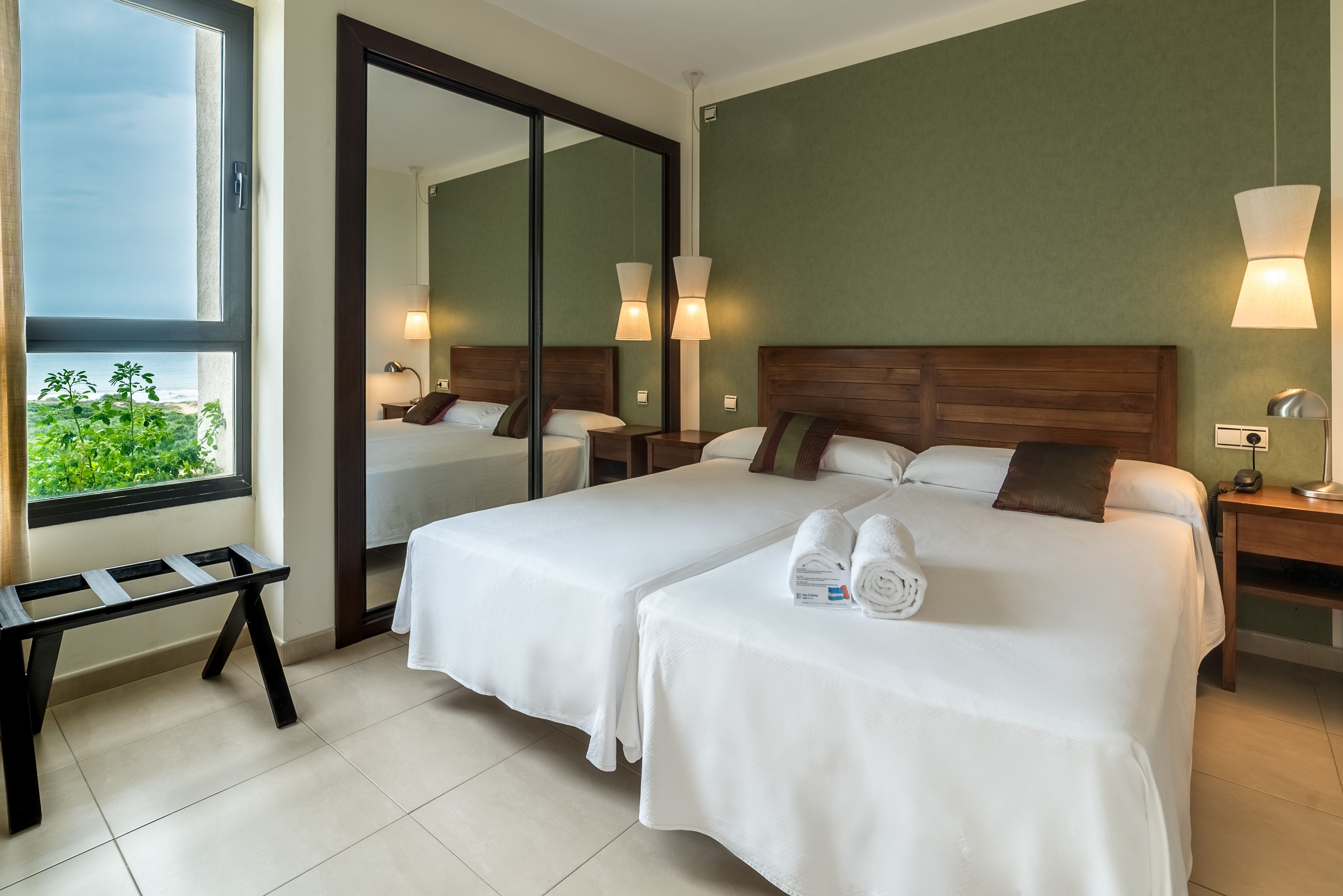 A hotel room with two beds, a large mirror, a bench, and a view of greenery outside.