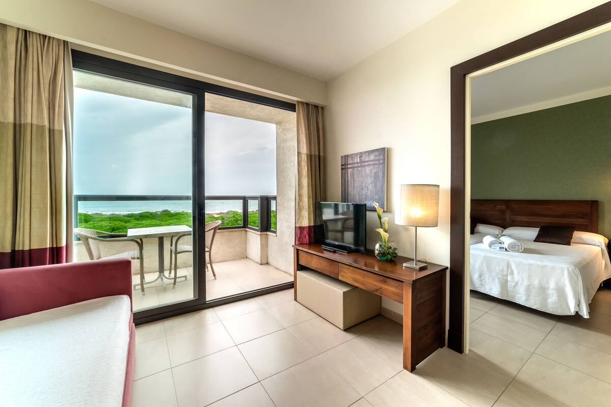 Een hotelkamer met balkon, bureau met televisie, bed en uitzicht op zee.