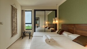 Habitación de hotel con dos camas, un espejo grande, vista a zonas verdes y una pequeña mesa con una silla.