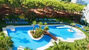 Una gran piscina de varios niveles con un puente que conecta diferentes zonas, rodeada de palmeras y sillas de descanso.