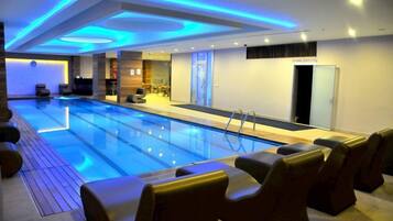 En moderne indendørs swimmingpool med blå belysning, et reception område og siddepladser langs poolen.