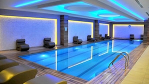 A modern indoor swimming pool with blue lighting, multiple seating areas, and a wooden deck. - Ozpark Hotel (Aksehir)