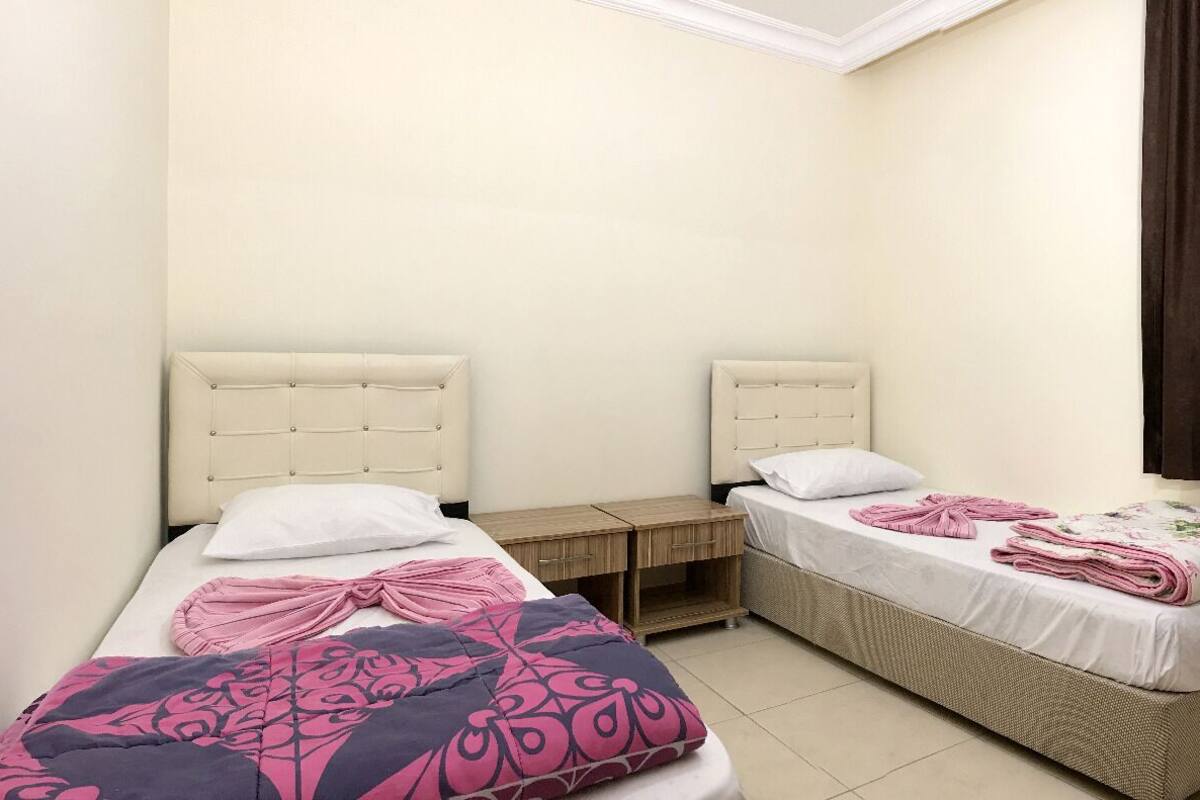 A small, clean room with two beds, a wooden nightstand, and a window with curtains.