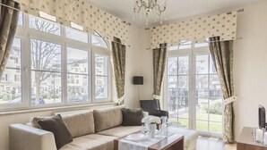 Un moderno salón con sofá, una mesa de centro y un mueble para la televisión. Amplias ventanas con cortinas, una lámpara de araña y vistas a edificios exteriores.