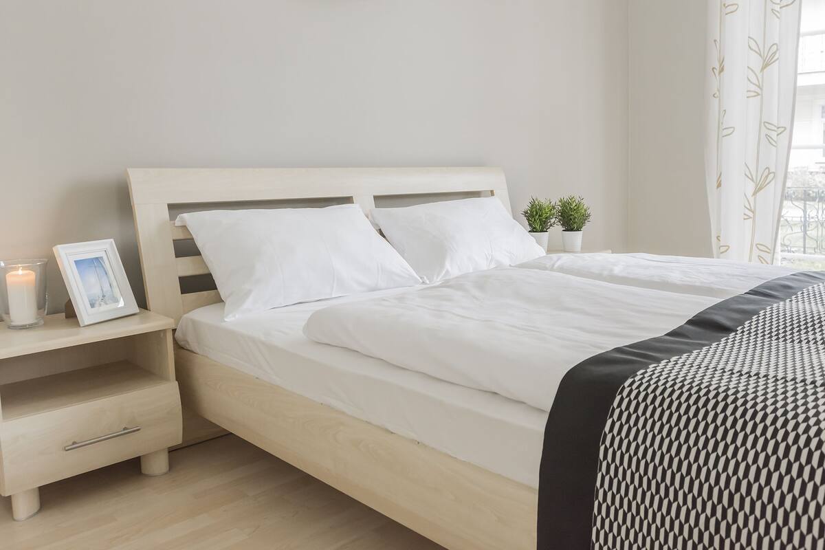Una cama bien hecha con ropa de cama blanca, una mesita de noche con una foto enmarcada y una vela, y una planta pequeña en la mesita de noche.