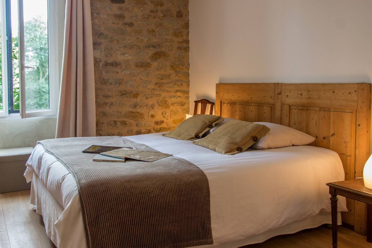 Une chambre avec un mur en pierre, une tête de lit en bois, un lit avec une literie blanche, une table de chevet en bois et une fenêtre avec des rideaux.