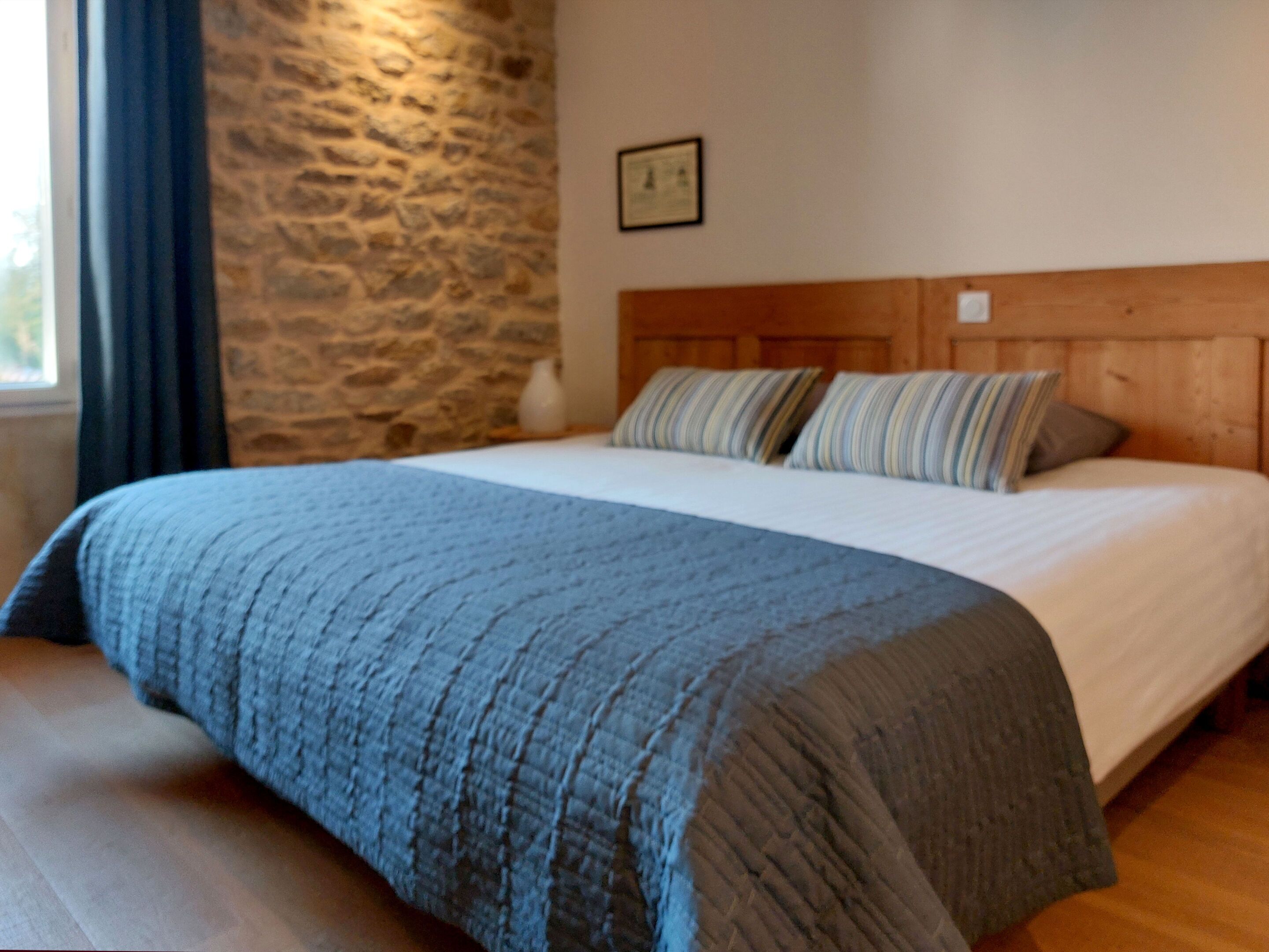 A bedroom with a stone wall, a wooden headboard, a bed with a blue blanket, and a bedside table with a vase.