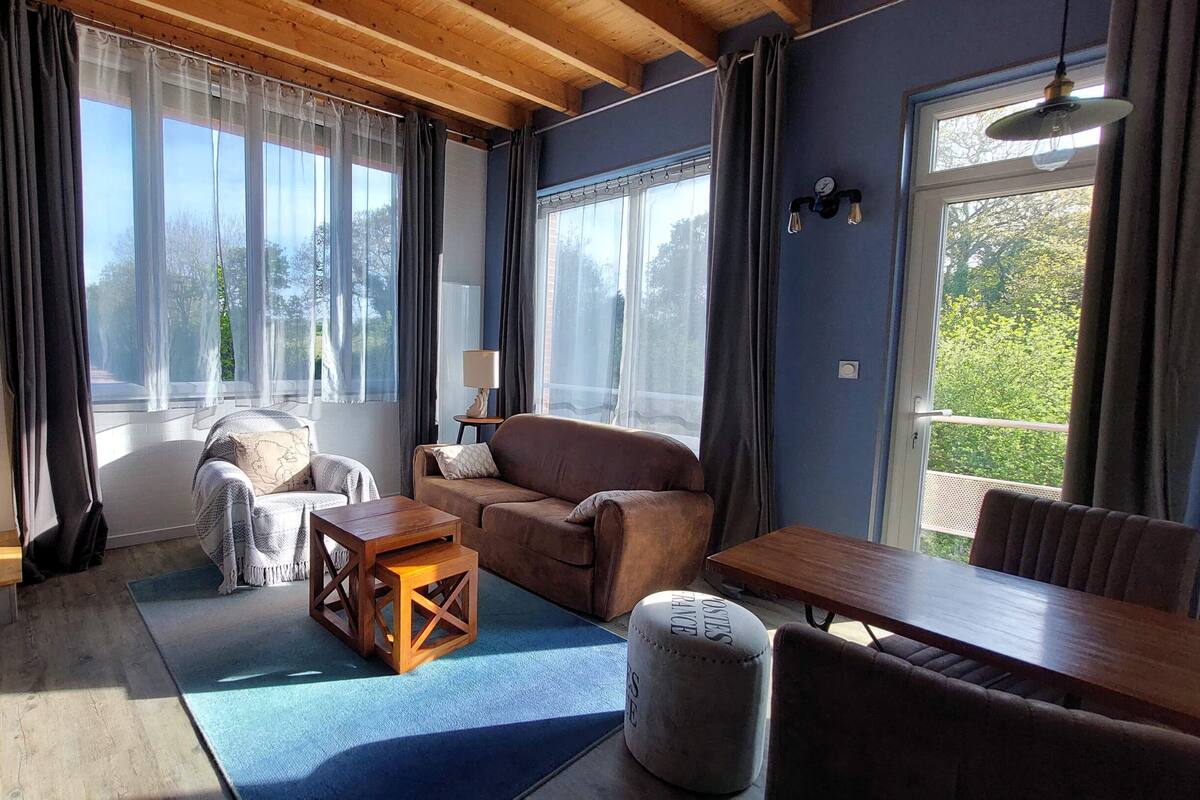 Un salon chaleureux avec un canapé, une table basse en bois et un petit pouf. De grandes fenêtres, ornées de rideaux, laissent entrer la lumière, et une lampe éclaire l’espace. Dehors, on aperçoit un paysage verdoyant.