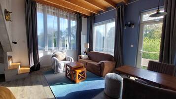 Un salon chaleureux avec un canapé, une table basse en bois et un petit pouf. De grandes fenêtres, ornées de rideaux, laissent entrer la lumière, et une lampe éclaire l’espace. Dehors, on aperçoit un paysage verdoyant.