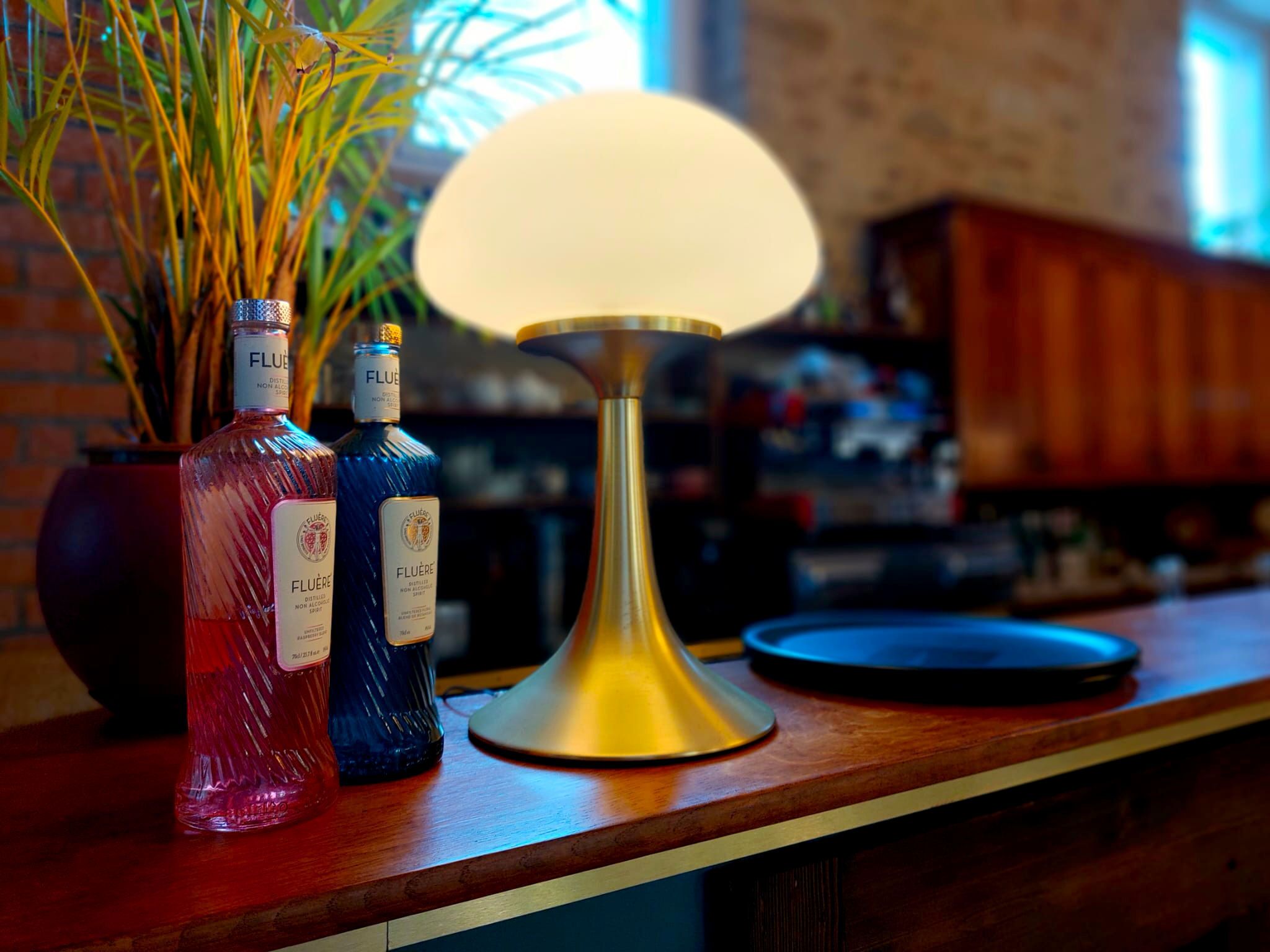 Un comptoir de bar avec deux bouteilles de FLUÈRE, une lampe de couleur dorée et une plante en pot.