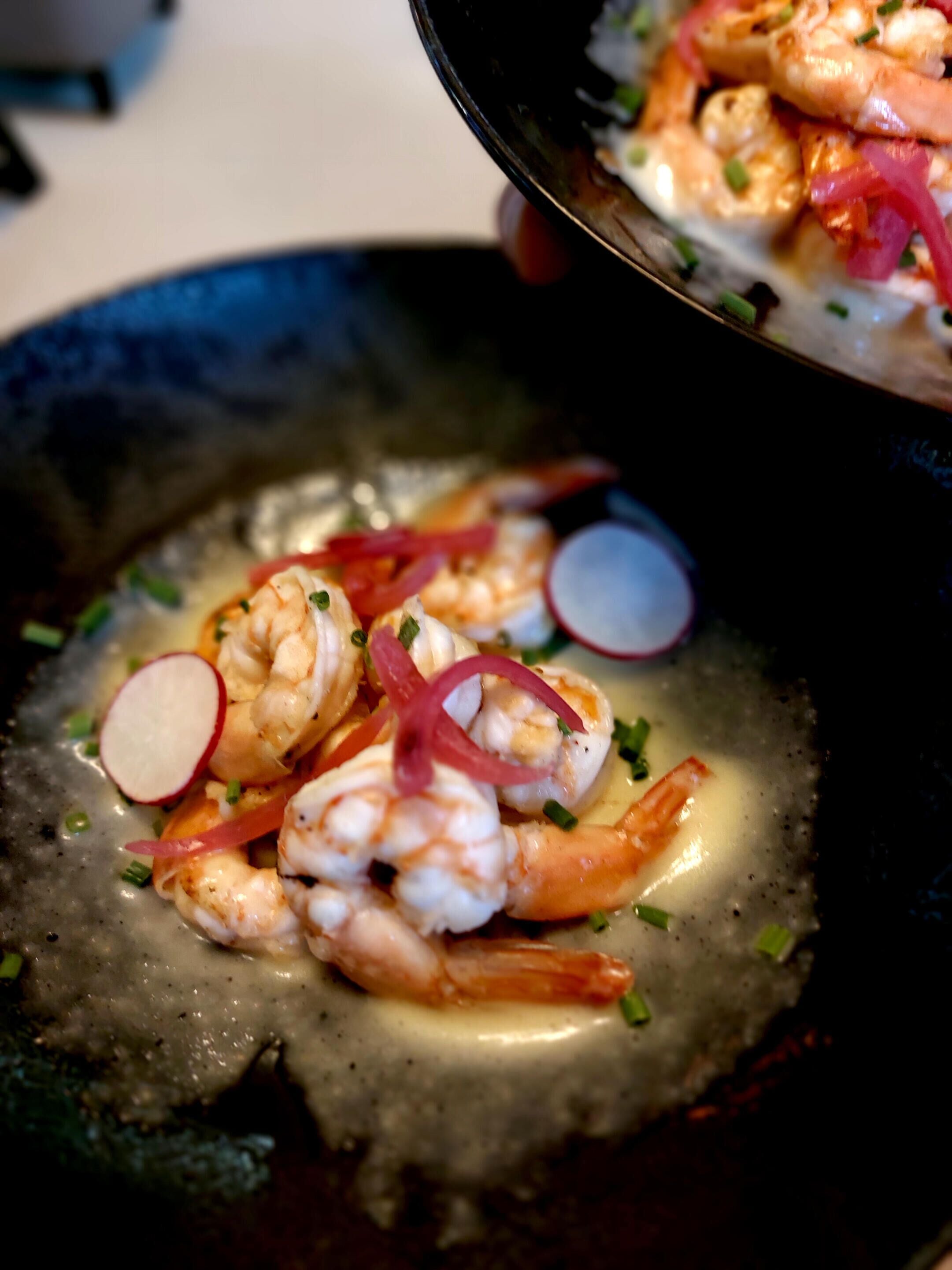 Un plat composé de crevettes, de fines tranches de radis et d’oignons verts, servi dans une assiette foncée.