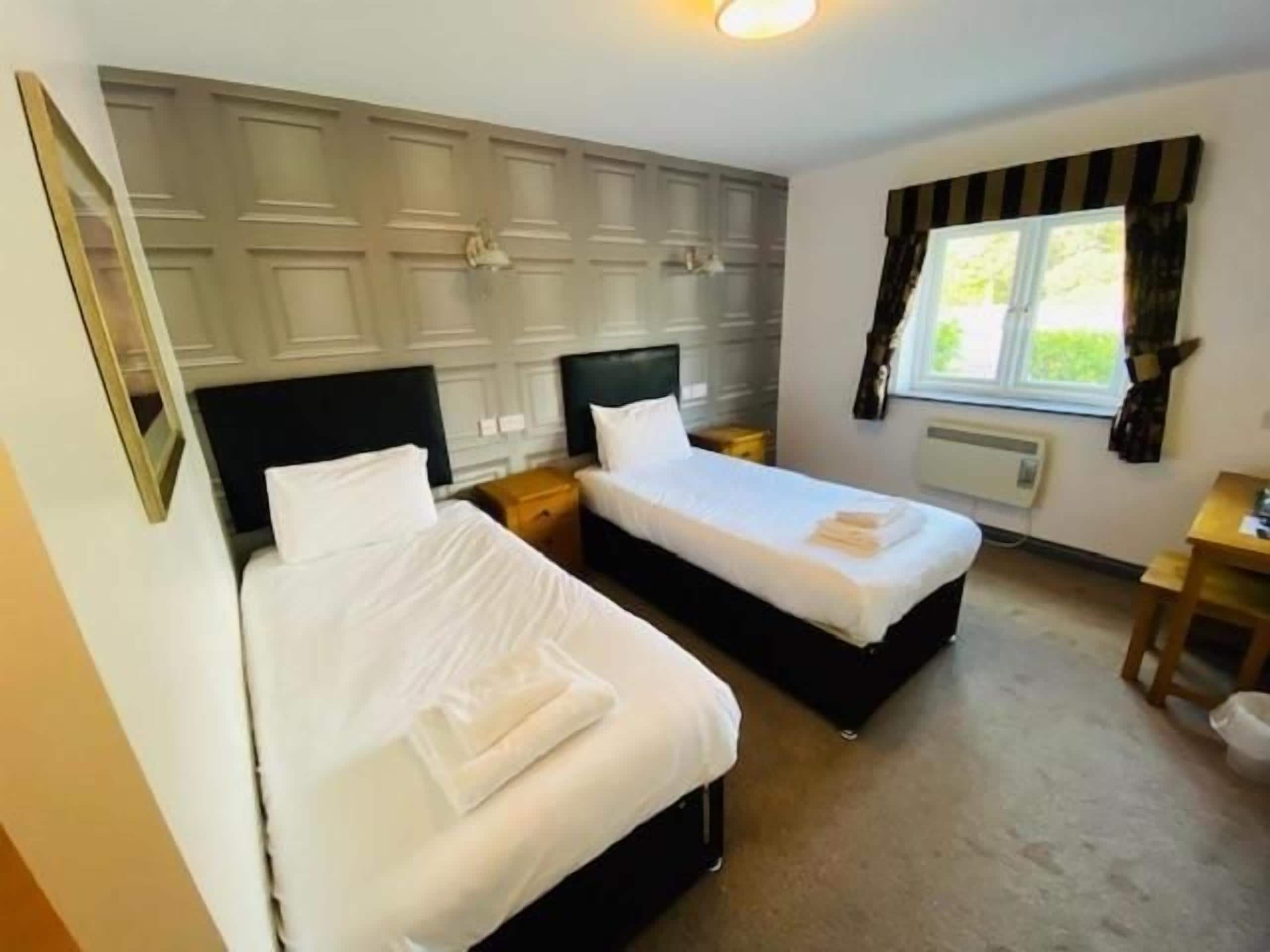 A hotel room with two single beds, a bedside table, a nightstand, a window with curtains, and a radiator.