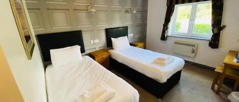 A hotel room with two single beds, a bedside table, a nightstand, a window with curtains, and a radiator.