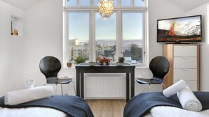 A modern room with a large window, a table with a decorative light, two chairs, a TV, and a bed with towels. - Alfred's Studios (Reykjavik)