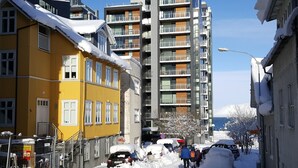 A snowy urban street with modern buildings, including a yellow building with a visible entrance and a multi-story building with balconies. - Alfred's Studios (Reykjavik)